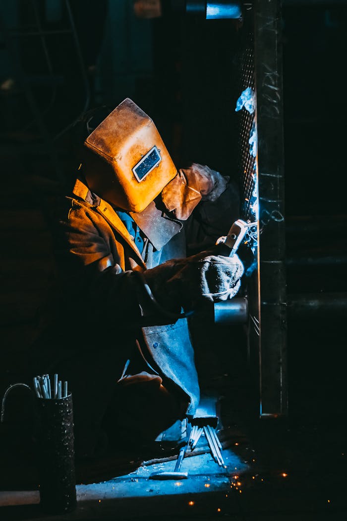 Home A welder wearing safety gear sparks metal in a dimly lit industrial setting.